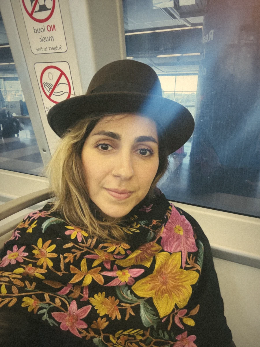 Saba smiling on a train, wearing a black hat and a black shawl embroidered with bright flowers
